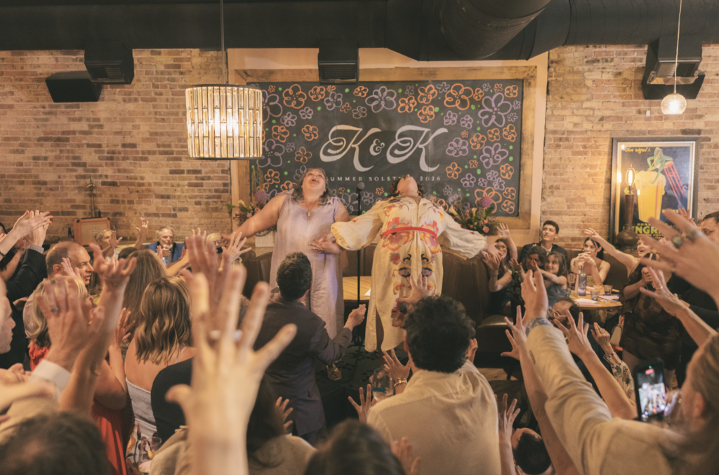 Krissy & Kate at their wedding getting sent energy by a crowd of attendees in front of a chalkboard sign beautifully decorated by Ash & Laurea